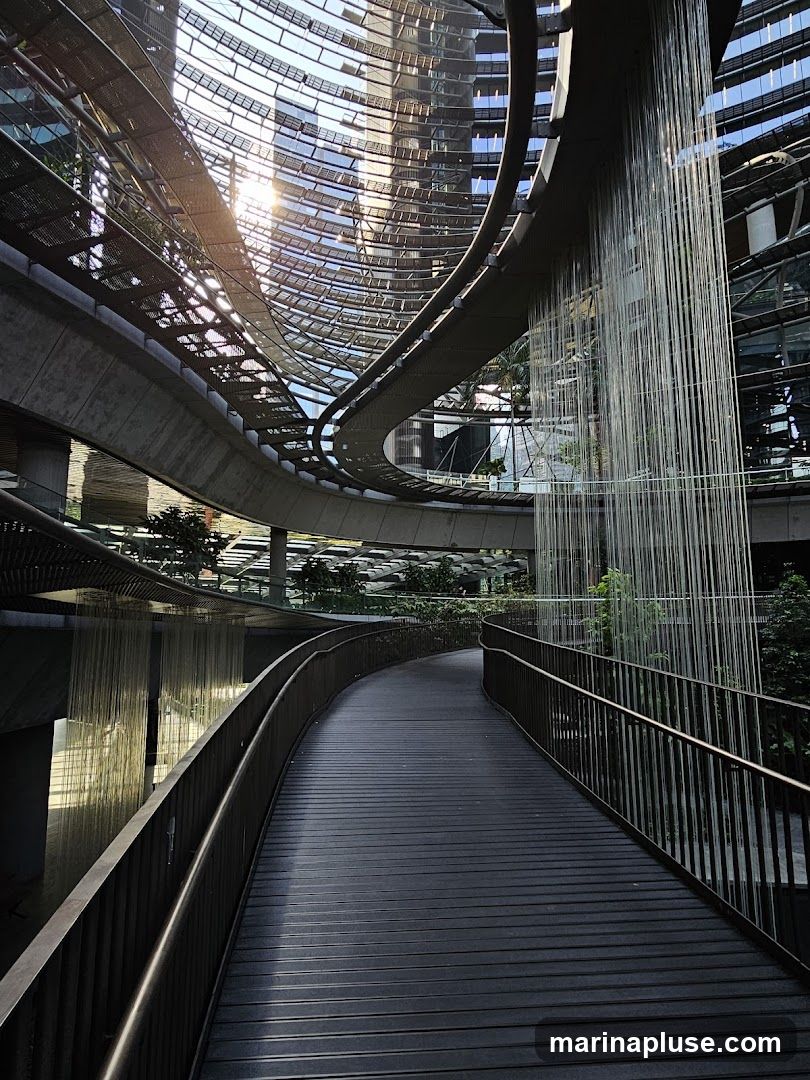 A dark, slatted walkway curves through a modern architectural space. The walkway is bordered by a dark metal railing. Above, a curved, transparent roof structure allows sunlight to stream in, reflecting off the glass facades of surrounding buildings. A striking feature is a vertical cascade of thin, light-colored strands, resembling a waterfall, descending from an elevated walkway. Lush greenery is visible in the background, integrated into the architecture.
