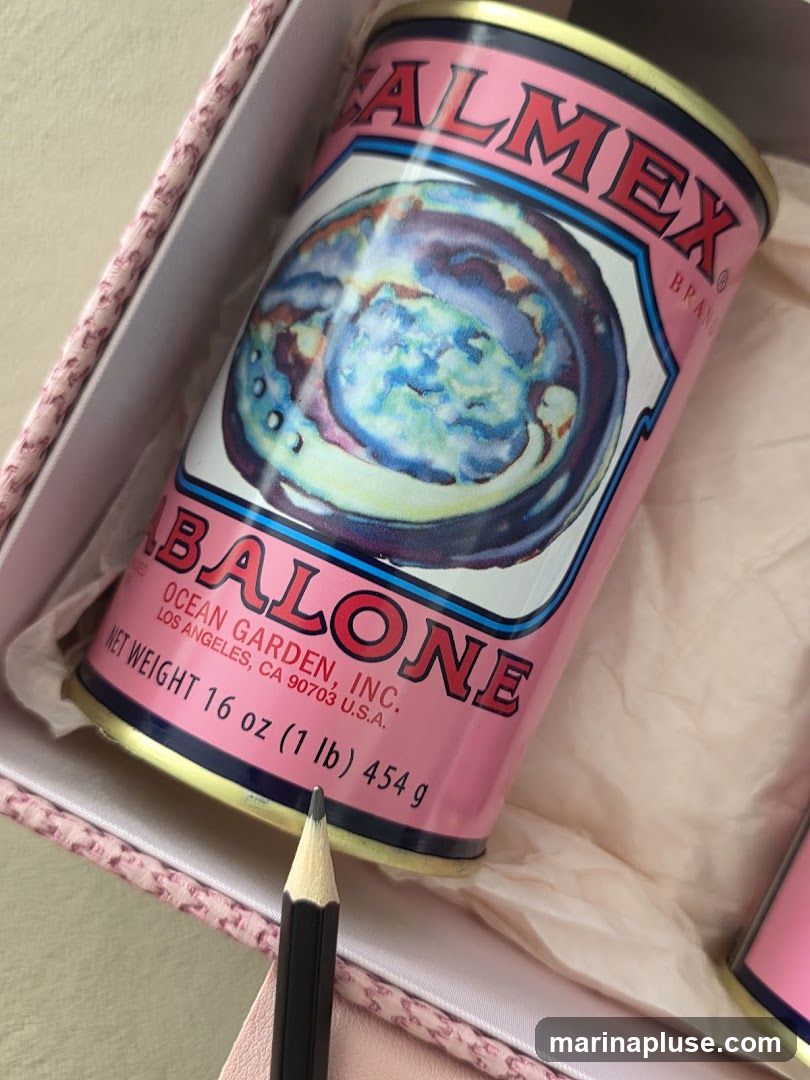 A close-up shot of a pink can of abalone, with a black pencil in the foreground. The can has a colorful illustration of abalone on it and text indicating the brand "ALMEX" and "OCEAN GARDEN, INC.". The net weight is listed as 16 oz (1 lb) 454 g. The can is placed inside a pink decorative box.