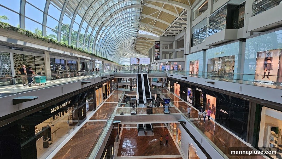 A wide-angle view of the interior of a multi-level shopping mall with a distinctive curved glass roof. Escalators are visible in the center, connecting different floors. Various luxury brand stores, such as Saint Laurent, Prada, and Miu Miu, are identifiable by their signage on the storefronts. People are seen walking on the elevated walkways and browsing in the shops. The mall features polished wooden floors and modern architectural elements.
