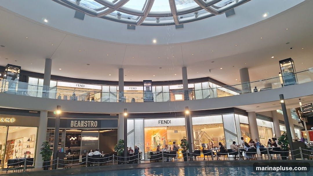 The image shows the interior of a multi-level shopping mall with a large, circular atrium. A water feature cascades from the ceiling into a pool below. Several high-end retail stores, including Fendi, are visible on the upper levels, with their prominent signage. On the lower level, a cafe or restaurant named 'BEANSTRO' is open, with patrons seated at tables. The overall ambiance is bright and contemporary, with a focus on luxury retail.