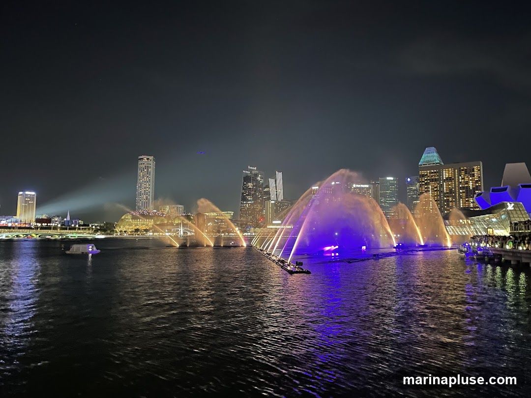 A nighttime view of a water fountain show with colorful lights, set against a backdrop of a city skyline. The water jets are illuminated with purple and orange lights, creating a dramatic display over the dark water. Several boats are visible on the water, and the city buildings are lit up, reflecting on the water's surface. The sky is dark, indicating it is nighttime.