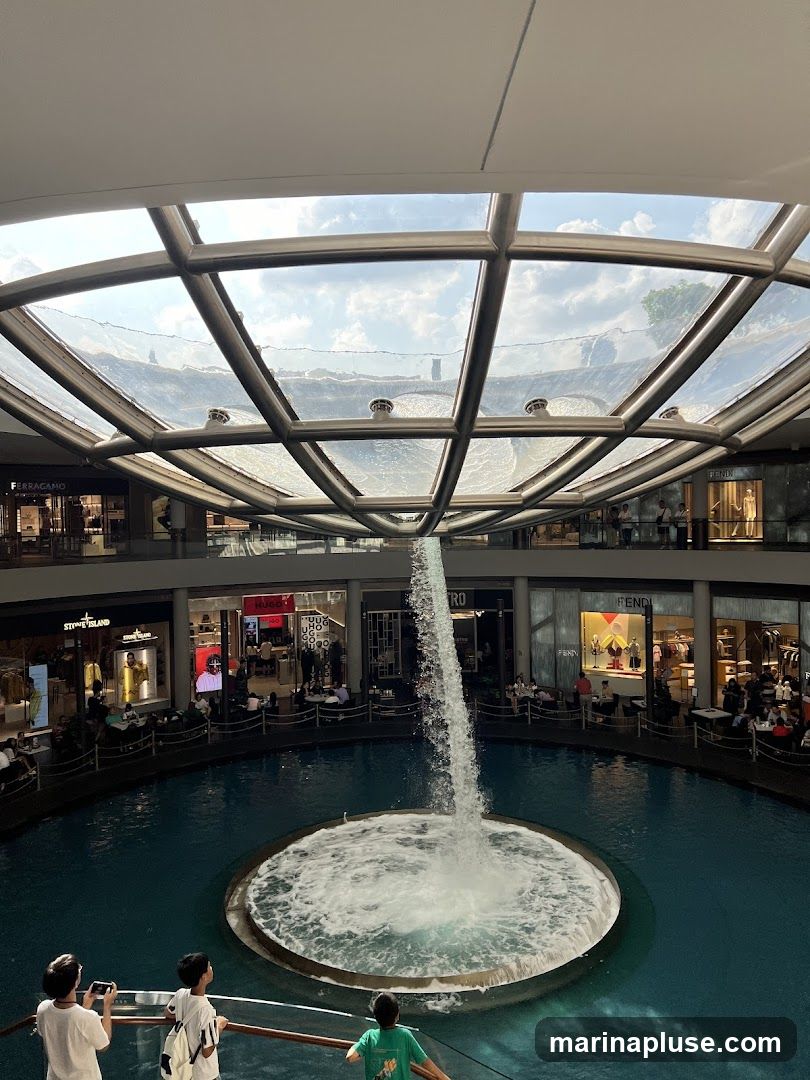 An interior view of a modern shopping mall featuring a large, circular water fountain with a stream of water cascading from above. The mall has multiple levels with various luxury brand stores visible, including Ferragamo, Fendi, and Stone Island. People are seen browsing in the shops and sitting around the water feature. The ceiling is a glass and metal structure, allowing natural light to filter in.