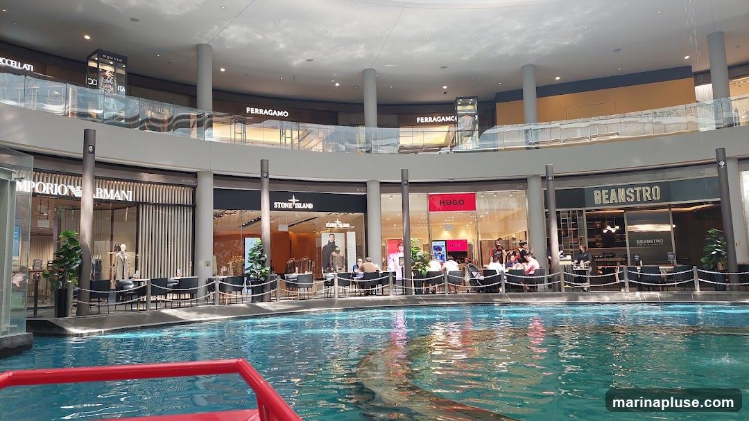 An interior view of a multi-level shopping mall with a canal or water feature in the foreground. Several high-end retail stores are visible on the ground level, including Emporio Armani, Stone Island, Hugo, and Beanstro. The upper levels also show storefronts with brand names like FERRAGAMO and MCQUEEN. People are seated at tables outside some of the establishments, suggesting a dining or lounge area. The architecture features a large, domed ceiling and numerous columns.