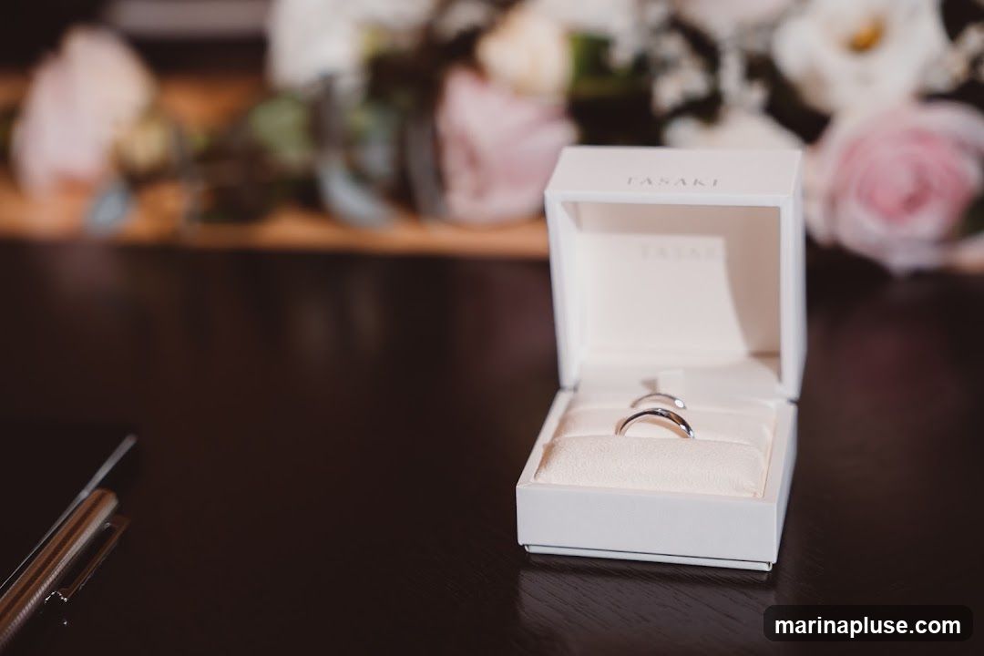 A white ring box with the brand name TASAKI embossed on it, containing two silver wedding bands. The box is open and sits on a dark wooden surface. In the blurred background, there are arrangements of pink and white flowers. To the left of the box, a pen and a dark object, possibly a phone, are visible.