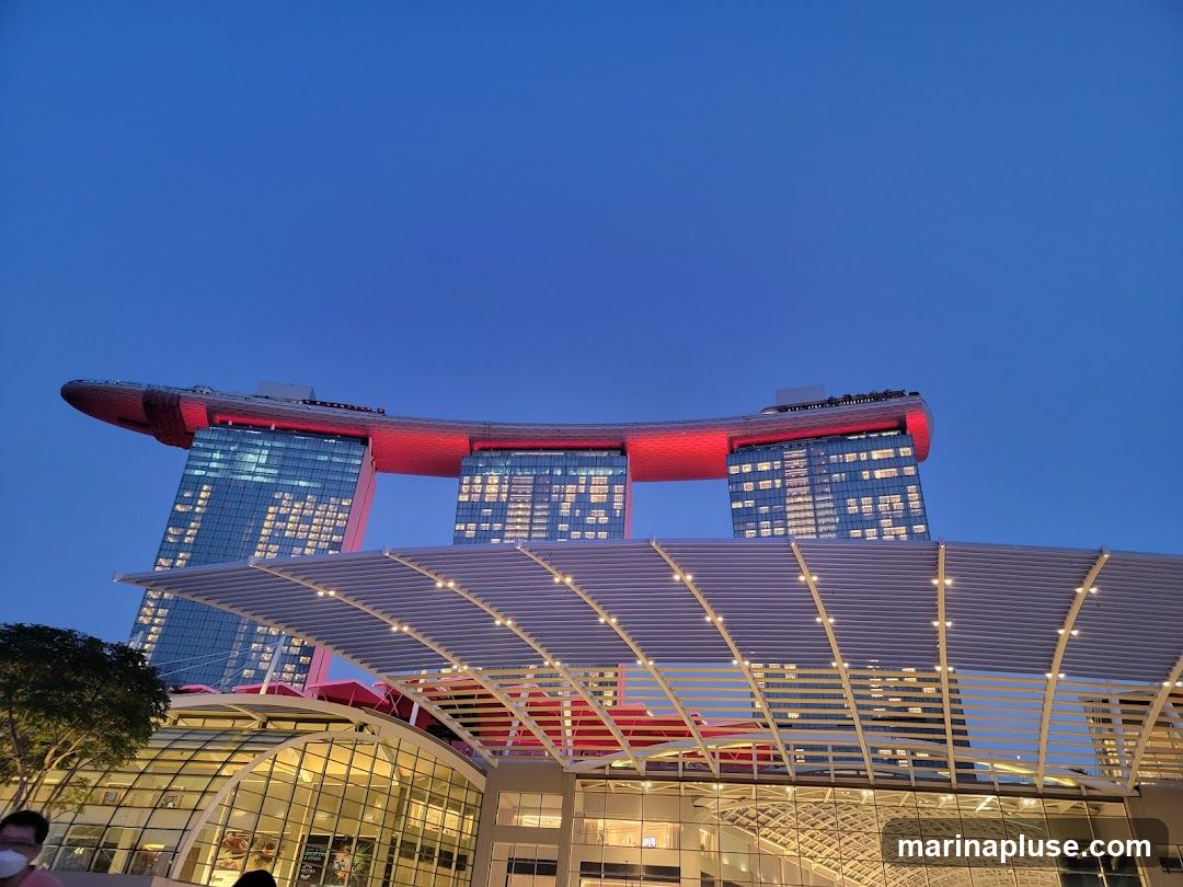 A wide shot of the Marina Bay Sands complex in Singapore as dusk settles. The iconic three towers are visible, topped with the red Sands SkyPark. The foreground features a modern, glass-covered structure with a white, slatted roof. The sky is a deep blue, transitioning from the day's light.