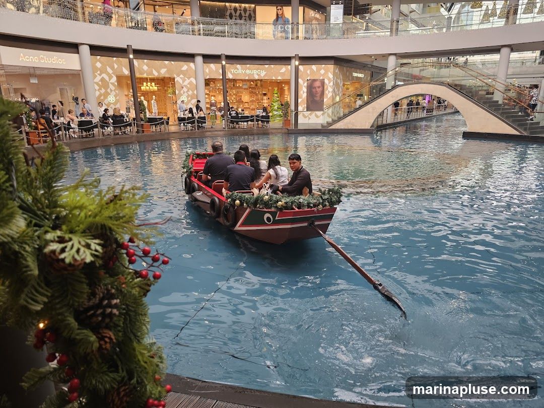 A boat with several passengers is being rowed through a canal inside a large, multi-level shopping mall. Shops like Acne Studios and Tory Burch are visible on the upper levels, along with escalators and bridges. A festive garland with pinecones and red berries is in the foreground.