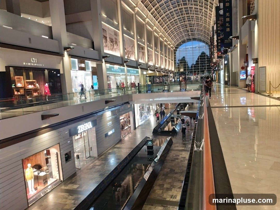 A view from an upper level of a spacious, multi-level shopping mall with a glass ceiling. Several storefronts are visible on both levels, including brands like Zegna and La Mer. People are seen walking around and browsing. The mall features reflective floors and a central water feature or channel.