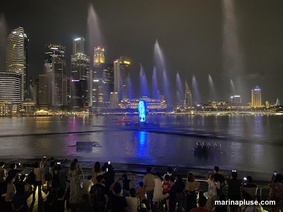 A crowd of people watches a water and light show at night, with a city skyline in the background. The show features numerous water jets illuminated with blue and white lights, creating a dramatic effect over the water. Several boats are visible on the water, and the city skyline is illuminated with many lights from tall buildings.