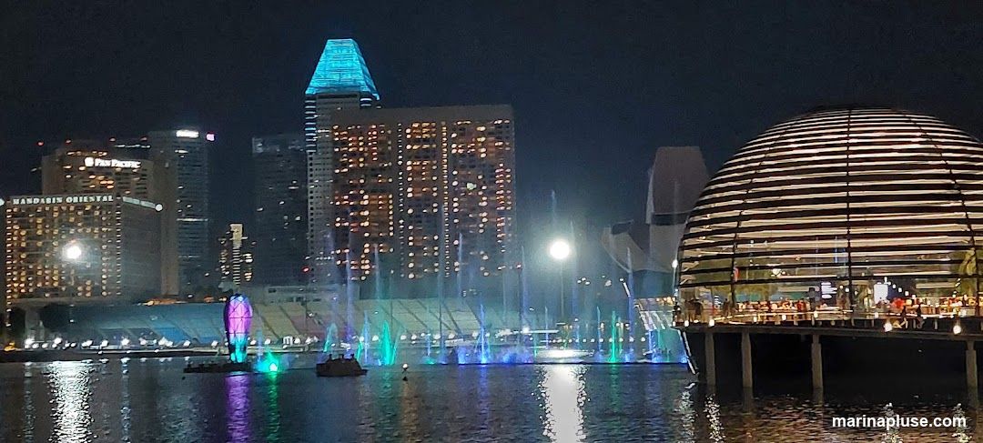 A nighttime view of a city skyline featuring illuminated skyscrapers and a domed building. In the foreground, a body of water reflects the lights, with a water and light show in progress. Several boats are visible on the water. The buildings include the Mandarin Oriental and Pan Pacific hotels.