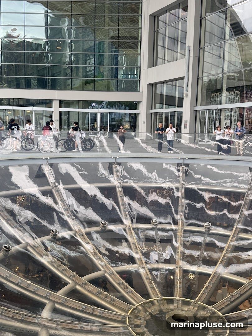 A wide shot of a modern building with large, reflective glass windows. In the foreground, a circular fountain structure with metallic supports is visible. People are seen standing on a walkway in front of the building, some with bicycles. The reflection in the windows shows a "The World's Studio Ghibli" sign and other architectural details.
