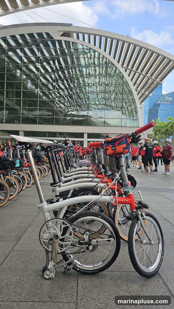 A row of folded bicycles, primarily Brompton brand, are parked on a paved plaza in front of a large, modern building with a distinctive curved glass facade. The building's exterior reflects the sky and surrounding structures. Several people, some wearing red shirts, are gathered in the background, suggesting an organized event or group activity. The bicycles are various colors, with a prominent red one in the foreground.