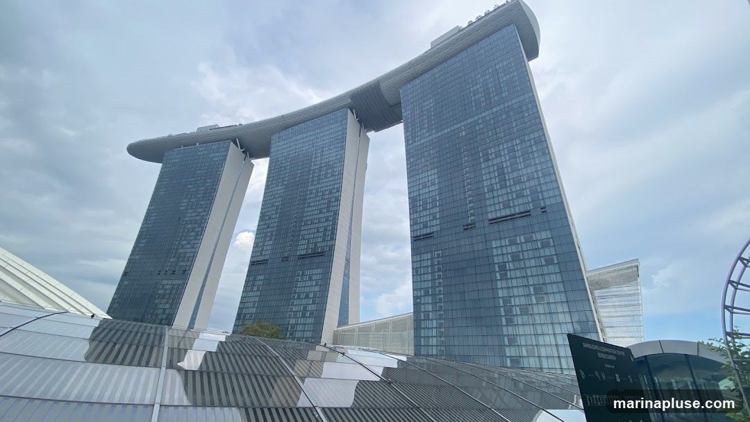 A wide-angle shot of the Marina Bay Sands hotel in Singapore, with its distinctive three towers connected by a sky-high platform. The sky is overcast with clouds. In the foreground, a modern, curved glass and metal structure is visible. A dark sign with white text, likely informational, is partially visible on the right side.