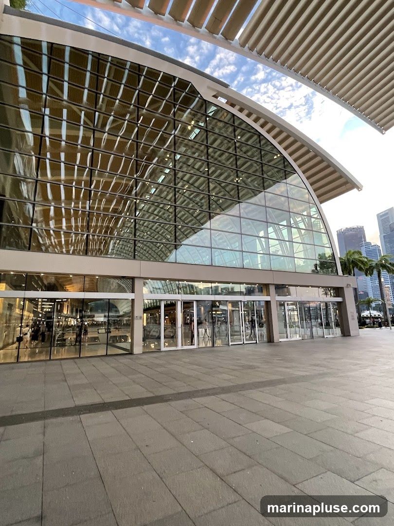A wide shot of a contemporary building with a large glass facade and a curved roof structure. The exterior features multiple glass doors and windows, reflecting the sky and surrounding architecture. The pavement in front is paved with large stone tiles.
