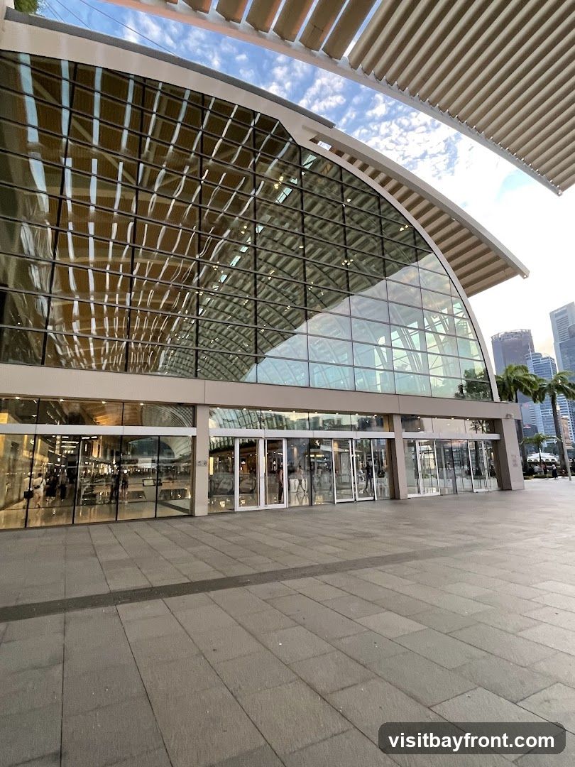 A wide shot of a contemporary building with a large glass facade and a curved roof structure. The exterior features multiple glass doors and windows, reflecting the sky and surrounding architecture. The pavement in front is paved with large stone tiles.