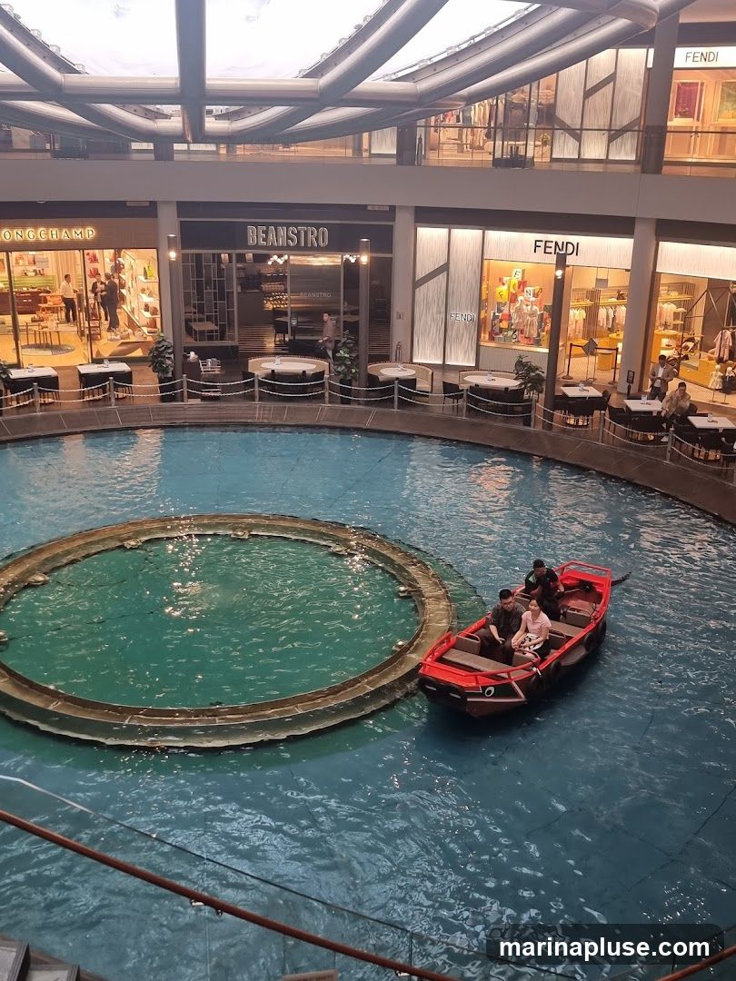An indoor canal with a red boat carrying passengers navigates through a shopping mall. Surrounding the canal are various shops, including Longchamp, Beanstro, and Fendi, with outdoor seating areas for diners. The mall features a modern architectural design with a large, open ceiling structure.