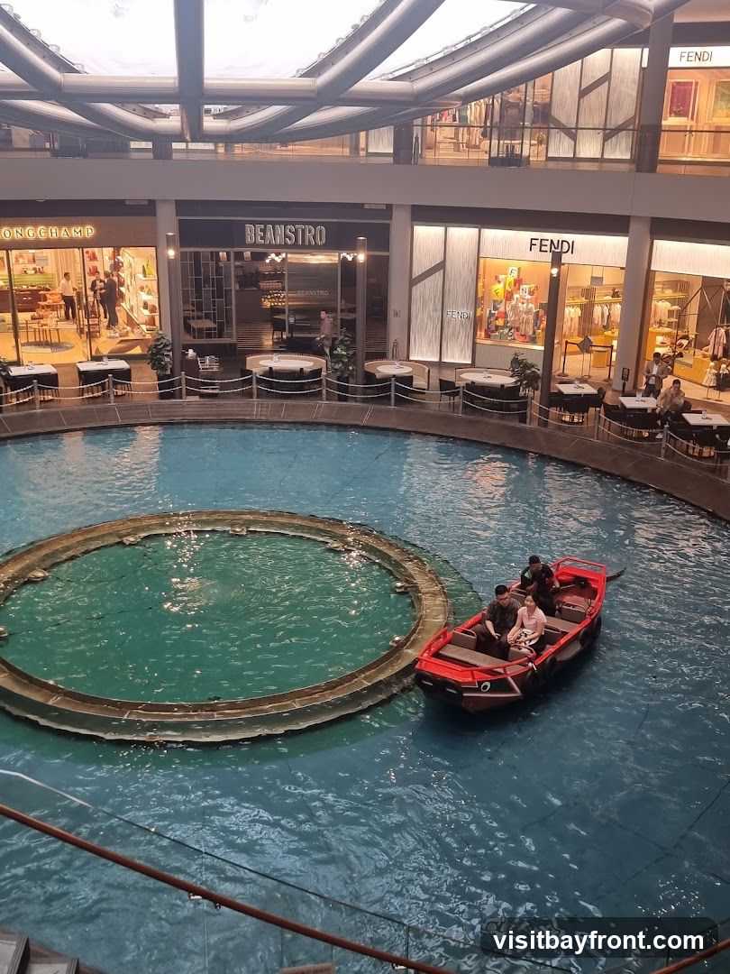 An indoor canal with a red boat carrying passengers navigates through a shopping mall. Surrounding the canal are various shops, including Longchamp, Beanstro, and Fendi, with outdoor seating areas for diners. The mall features a modern architectural design with a large, open ceiling structure.