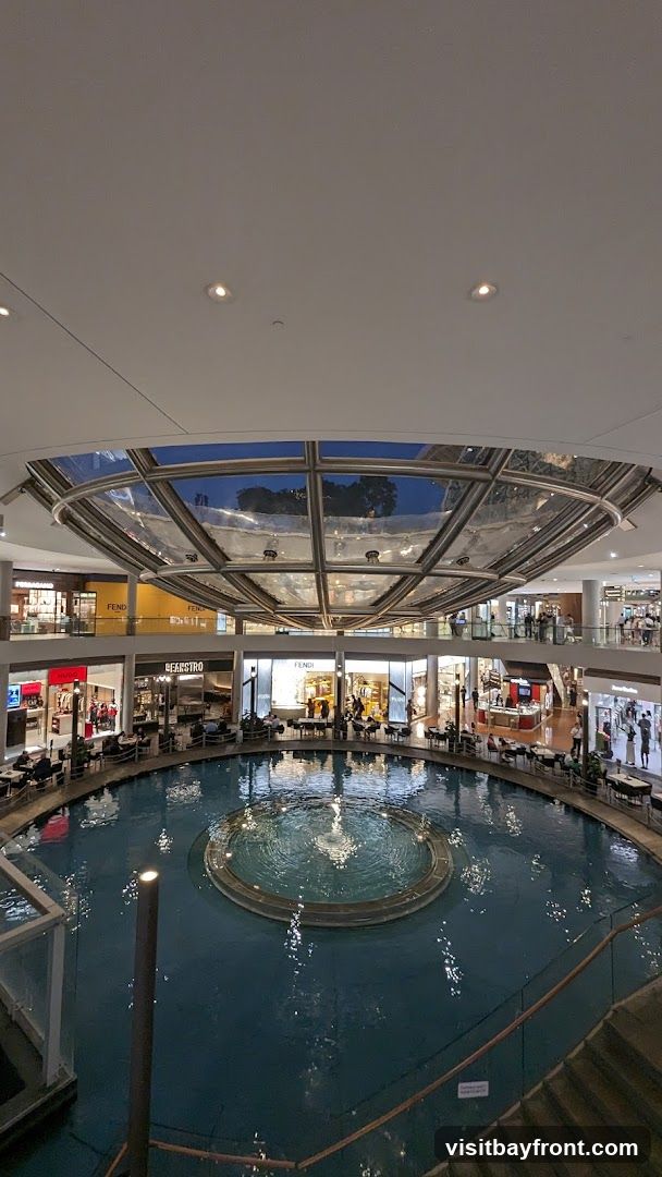 An interior view of a multi-level shopping mall featuring a large circular indoor water feature with a fountain. Shops and restaurants with seating areas surround the water feature. The mall has a modern design with a glass ceiling.