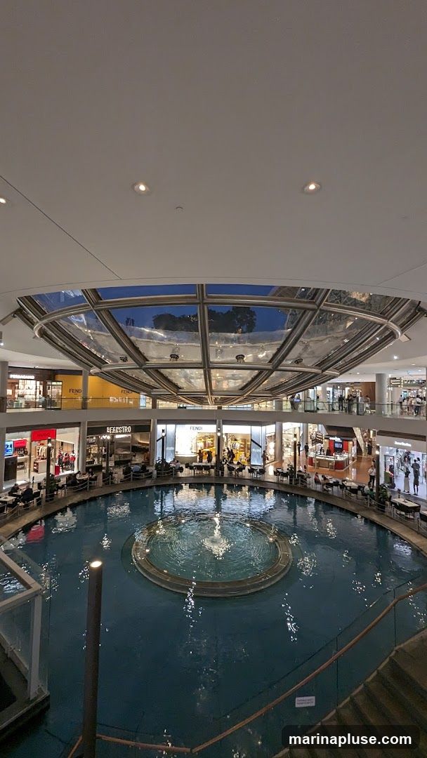 An interior view of a multi-level shopping mall featuring a large circular indoor water feature with a fountain. Shops and restaurants with seating areas surround the water feature. The mall has a modern design with a glass ceiling.