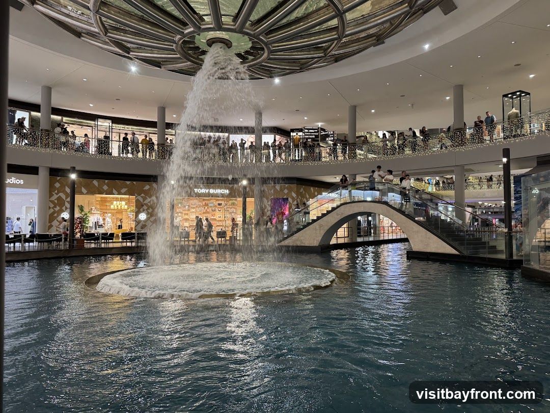 An indoor shopping mall features a large water feature with a fountain in the center. People are seen on multiple levels of the mall, looking at the fountain and browsing shops. A bridge connects two sides of the mall over the water. Stores like Tory Burch are visible.