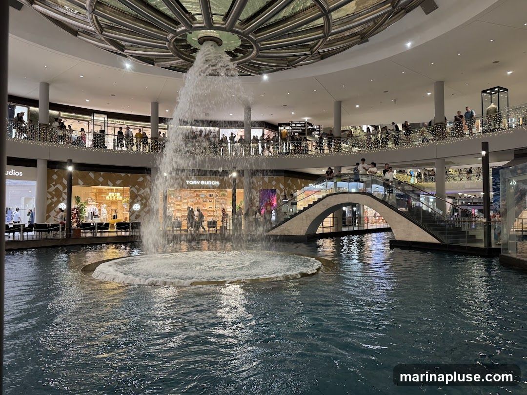 An indoor shopping mall features a large water feature with a fountain in the center. People are seen on multiple levels of the mall, looking at the fountain and browsing shops. A bridge connects two sides of the mall over the water. Stores like Tory Burch are visible.