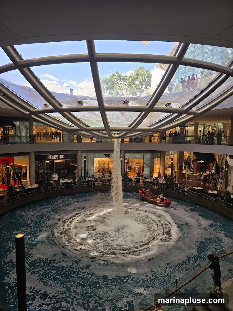 An indoor shopping mall with a large, circular water feature in the center. A fountain is active in the middle of the water, and a small boat is navigating the water. Shops with visible signage like HUGO, BEANSTR, FENDI, and OELIN are located on multiple levels surrounding the water feature. People are seen browsing in the shops and sitting at cafes. The mall has a modern design with a glass roof that allows natural light to enter.