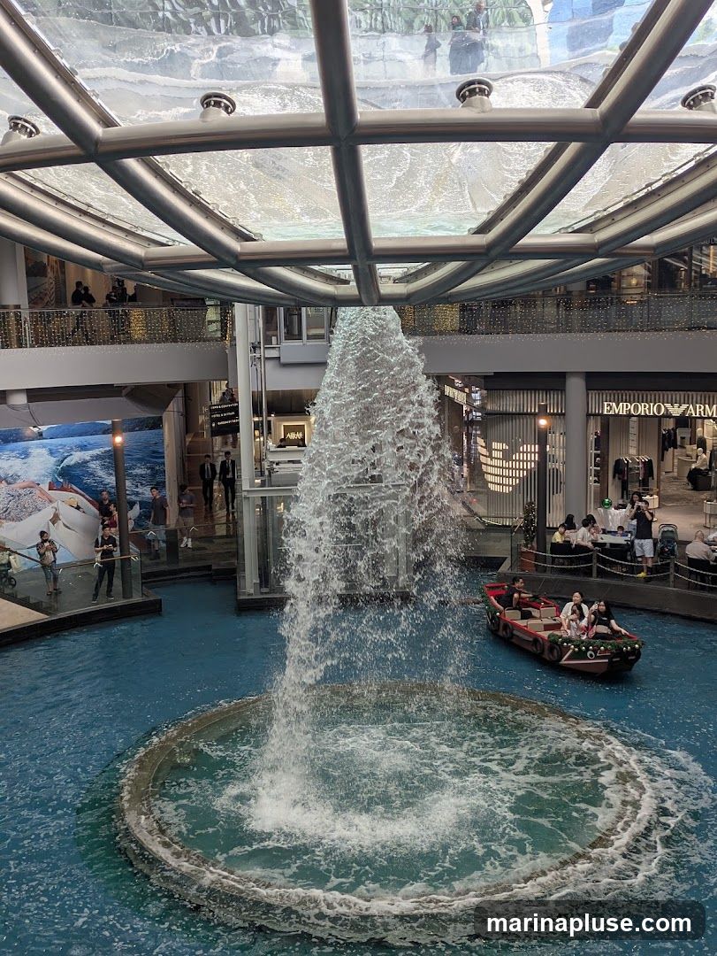 An indoor shopping mall features a large fountain in the center of a blue pool. A small boat with several passengers is navigating the water. Shops are visible on the upper levels, including one with an "EMPORIO ARMANI" sign. The ceiling is a glass and metal structure.