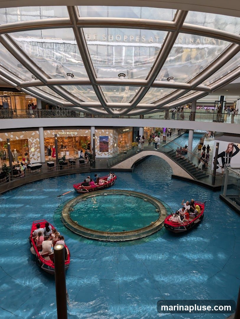 An indoor shopping mall features a canal with gondola-like boats carrying passengers. The canal is surrounded by multiple levels of shops, including visible storefronts for Gucci and Tory Burch. A large, transparent, geodesic dome-like ceiling allows natural light to filter in. The overall ambiance is that of a luxurious and modern shopping destination.