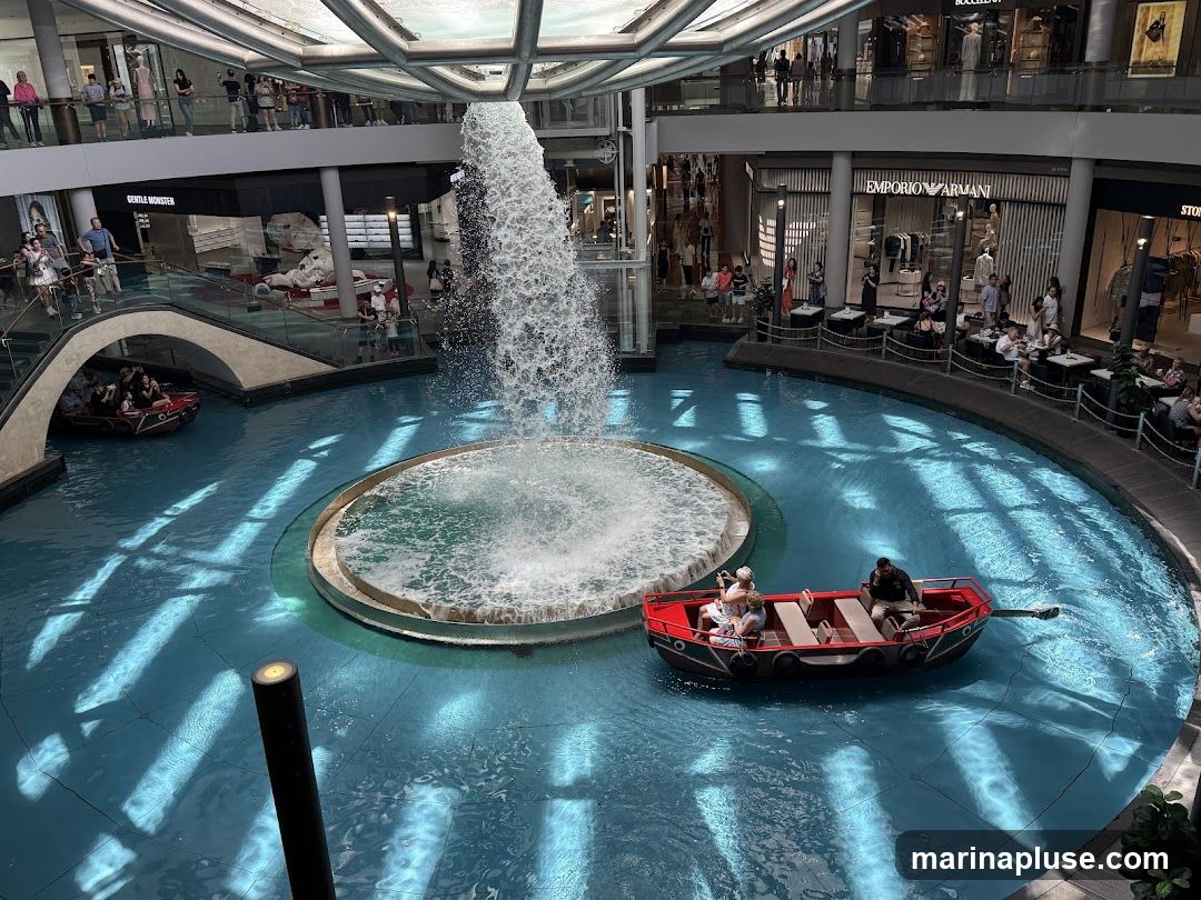 An indoor canal with clear blue water, featuring a large fountain in the center. Several small boats, some with passengers, navigate the canal. Surrounding the canal are multiple levels of a shopping mall with various storefronts, including Emporio Armani, and people walking and dining.