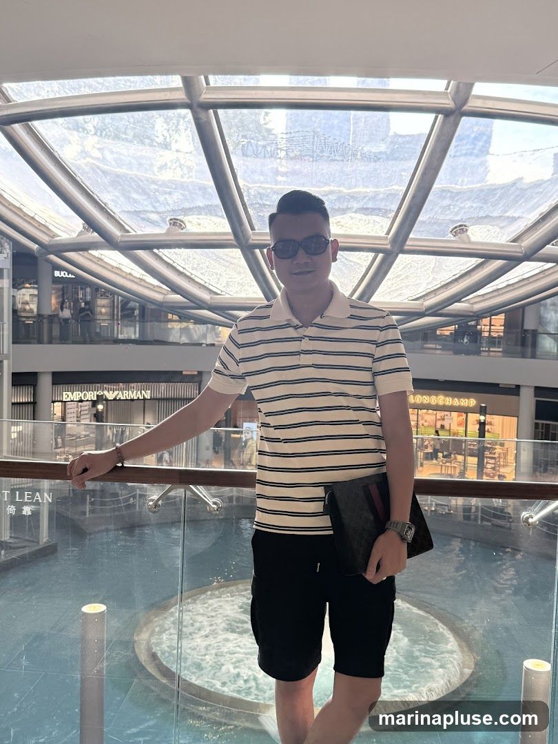 A man stands on a balcony overlooking a water feature in a modern shopping mall. He is wearing a striped polo shirt, black shorts, and sunglasses. He is holding a dark clutch bag. In the background, storefronts are visible, including Emporio Armani and Longchamp. The mall has a large glass ceiling structure.