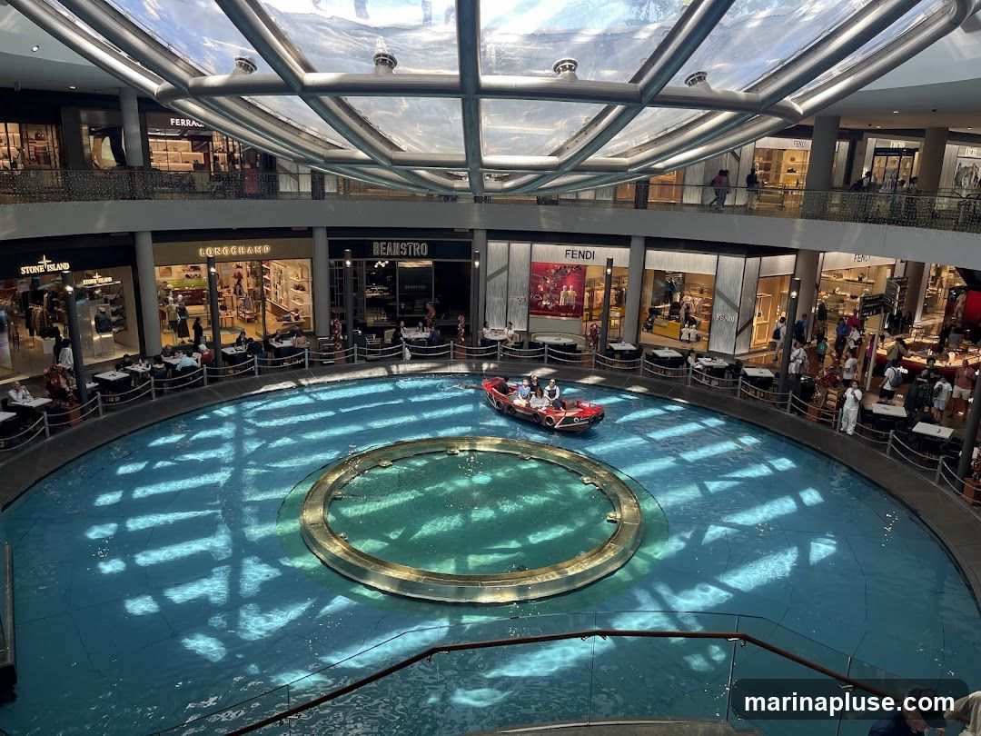 An interior view of a large, multi-level shopping mall with a canal running through the center. A small boat with people is navigating the canal. Shops such as Fendi, Longchamp, Stone Island, and Beanstro are visible on the ground floor, with tables and chairs set up outside some of them. The mall has a modern design with a glass ceiling that allows natural light to filter in, creating patterns on the water.