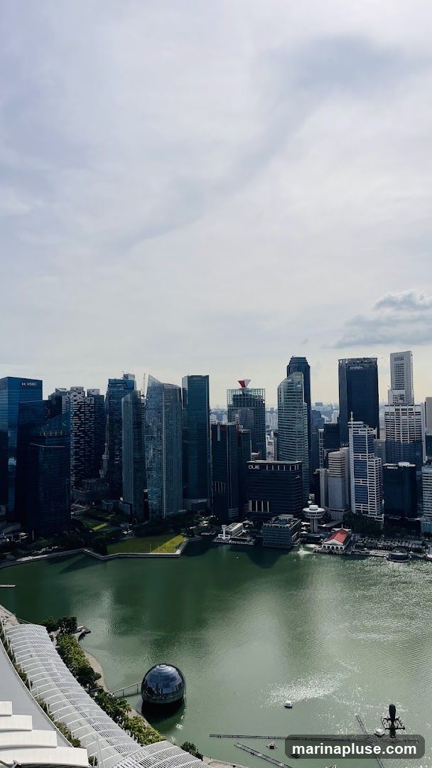 A panoramic view of the Singapore skyline, featuring numerous modern skyscrapers and the Marina Bay area. The water in the bay reflects the sky and buildings. A distinctive spherical structure is visible in the water, and a modern building with a white, curved roof is in the foreground. The sky is partly cloudy.
