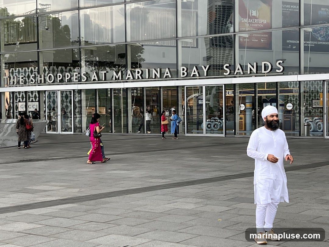 A wide shot of the exterior of The Shoppes at Marina Bay Sands. The building has a modern glass facade with large windows. The name of the mall is prominently displayed in large white letters above the entrance. Several people are walking in front of the building. A man in traditional Sikh attire is standing in the foreground. Signs for Gucci are visible on the storefront. Advertisements for Sneakertopia and Future World are displayed on the upper windows.