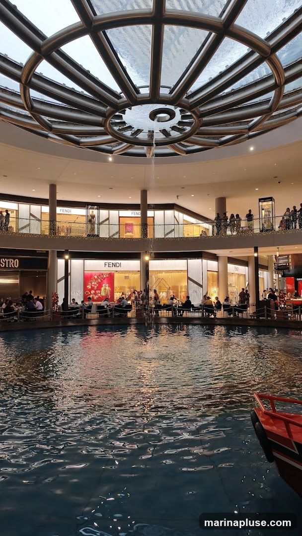 An indoor shopping mall with a large water feature in the foreground. The water reflects the lights and architecture of the mall. In the background, there are multiple levels of shops, including a Fendi store. People are seated at tables by the water, and there is a small red boat docked on the right side. The ceiling features a large, circular skylight with a water cascade.