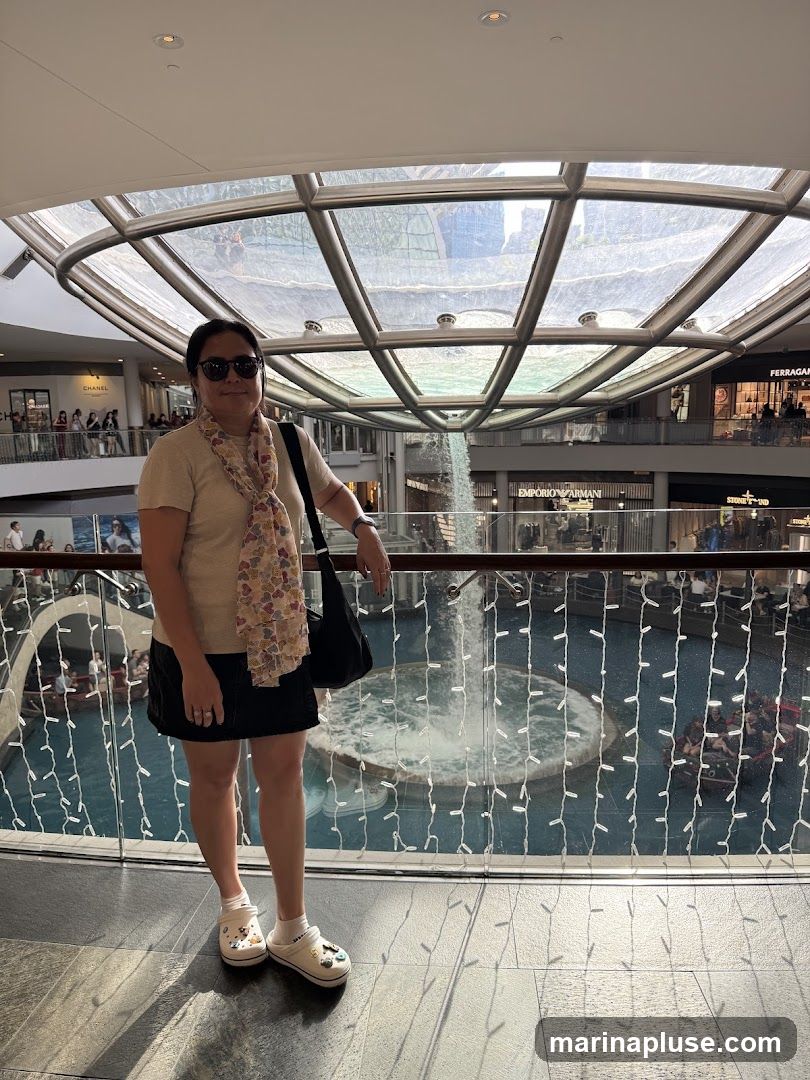 A woman stands on an upper level of a modern indoor mall, posing for a photo. She is wearing sunglasses, a light-colored short-sleeved shirt, a patterned scarf, and a black skirt. She has a black shoulder bag and is wearing white Crocs with colorful charms. In the background, a large indoor waterfall cascades into a pool surrounded by decorative lights. Shops with visible signage, including "CHANEL," "EMPORIO ARMANI," and "FERRAGAMO," are visible on the lower levels.