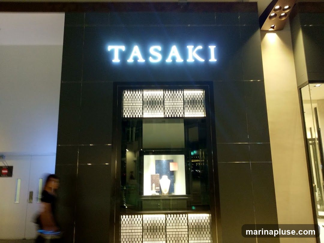 A well-lit storefront with the name "TASAKI" displayed in large, illuminated letters above a display window. The facade is dark and tiled, with decorative patterns above and below the window. Inside the window, jewelry is displayed on a stand. A person is walking past the entrance on the left side of the image.