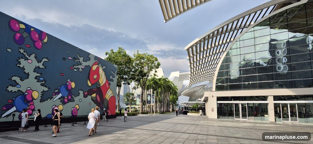 A wide shot of a modern building with a glass facade and an overhead canopy structure. To the left, a large mural featuring colorful, cartoonish figures covers a wall. Several people are walking on the paved plaza in front of the mural and the building. Palm trees line the walkway between the mural and the building. The sky is partly cloudy.
