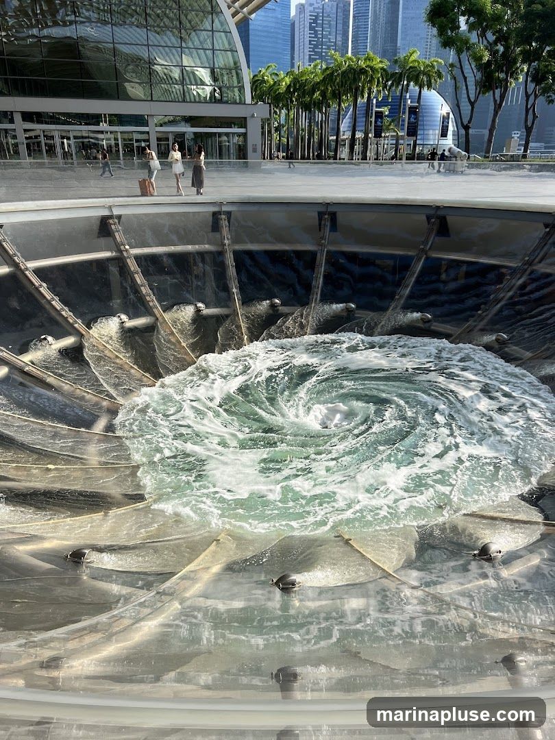 A close-up view of a large, circular water feature with a powerful vortex in the center. The water is churning and creating a whirlpool effect. In the background, a modern building with a glass facade and palm trees are visible, suggesting an urban outdoor setting.