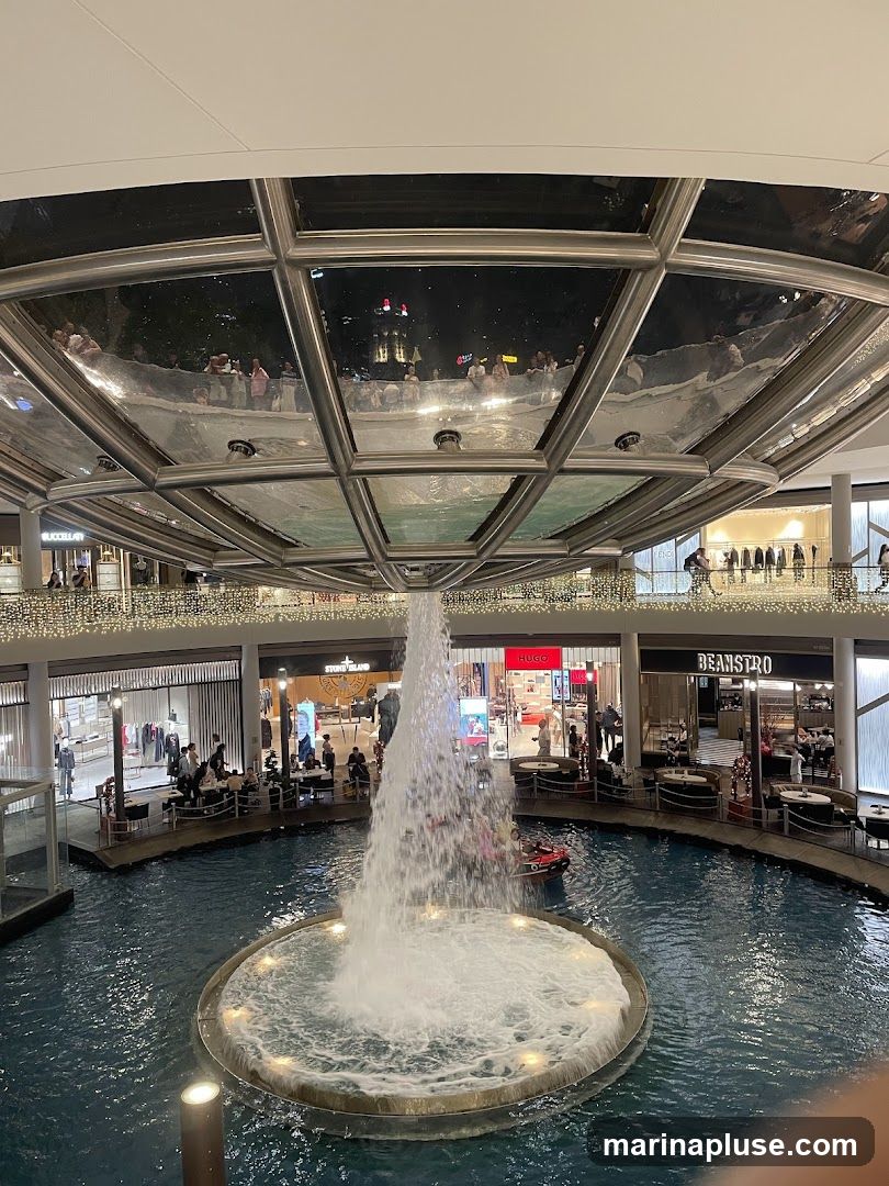 An indoor shopping mall features a large water fountain in the center, with shops and restaurants surrounding it. The ceiling has a reflective surface that mirrors the lights and people above. The shops visible include Hugo, Beanstro, and Stone Island.