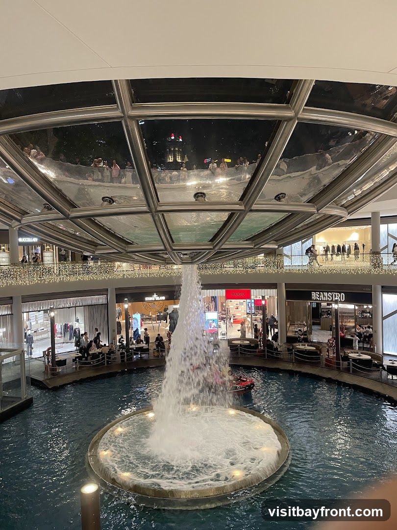 An indoor shopping mall features a large water fountain in the center, with shops and restaurants surrounding it. The ceiling has a reflective surface that mirrors the lights and people above. The shops visible include Hugo, Beanstro, and Stone Island.