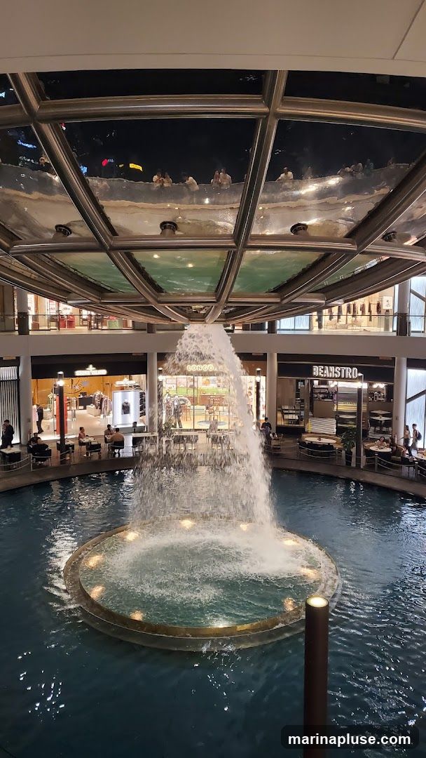 An indoor fountain with cascading water is the central feature of this scene. Surrounding the fountain are various shops and seating areas. One shop is labeled "BEANSTRO". Another shop has clothing displayed. People are seen dining at tables and walking around. The ceiling features a reflective surface with a grid pattern, showing reflections of people and lights.