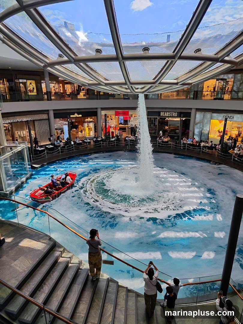An interior view of a modern shopping mall featuring a large, circular water feature with a powerful fountain in the center. A small red boat with several passengers navigates the water. Shops with visible signage such as FENDI, HUGO, STONE ISLAND, and BEANSTRO are located around the perimeter of the water feature. People are seen observing the water feature from a staircase in the foreground. The mall has a glass ceiling allowing natural light to enter.