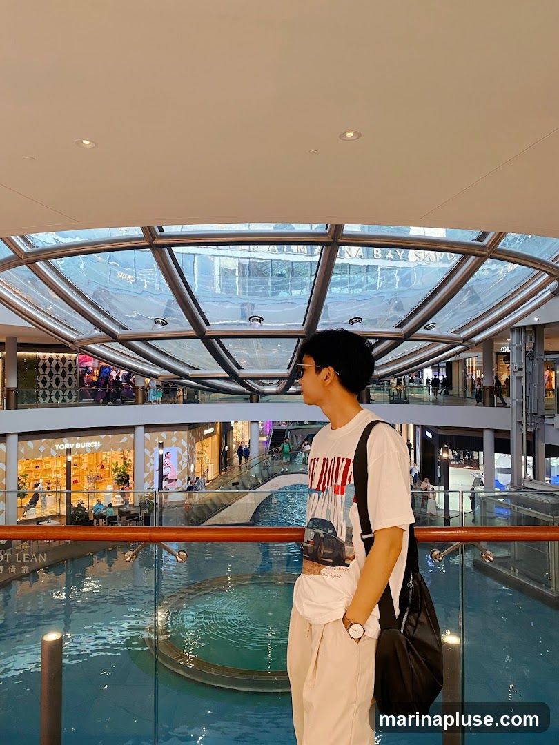 A young man stands on a balcony overlooking a large indoor water feature in a modern shopping mall. He is wearing a white t-shirt with a graphic print, light-colored pants, and a black shoulder bag. In the background, various shops are visible, including a Tory Burch store, and people are browsing and walking around. The mall features a large glass ceiling and multiple levels connected by escalators and walkways.