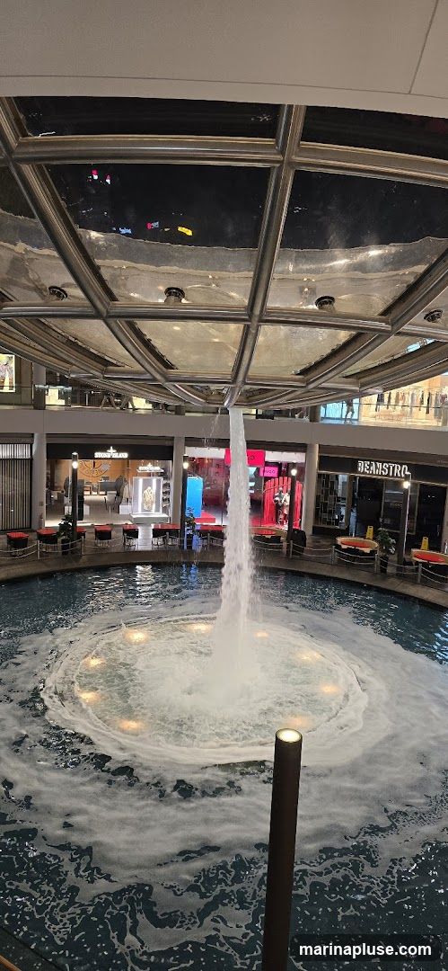An indoor fountain with a strong water jet is at the center of a circular pool. Surrounding the pool are various shops and seating areas. The shops have illuminated storefronts displaying merchandise. One shop has a prominent "STONE ISLAND" sign. Another visible sign reads "BEANSTRO". The ceiling is a glass and metal structure, allowing light to filter through and reflecting the interior.