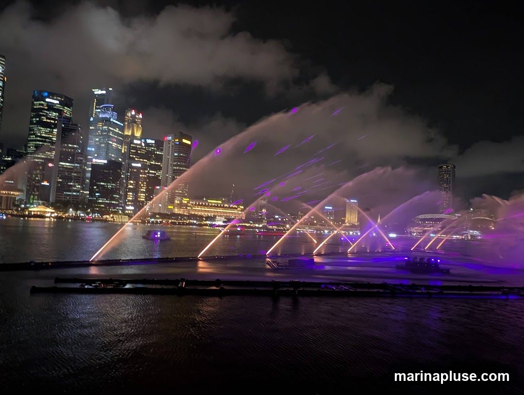 A vibrant water and light show takes place on a body of water at night, with jets of water illuminated by purple and orange lights. In the background, a modern city skyline with numerous illuminated skyscrapers stands against a cloudy night sky. Several boats are visible on the water, some appearing to be part of the show's infrastructure.