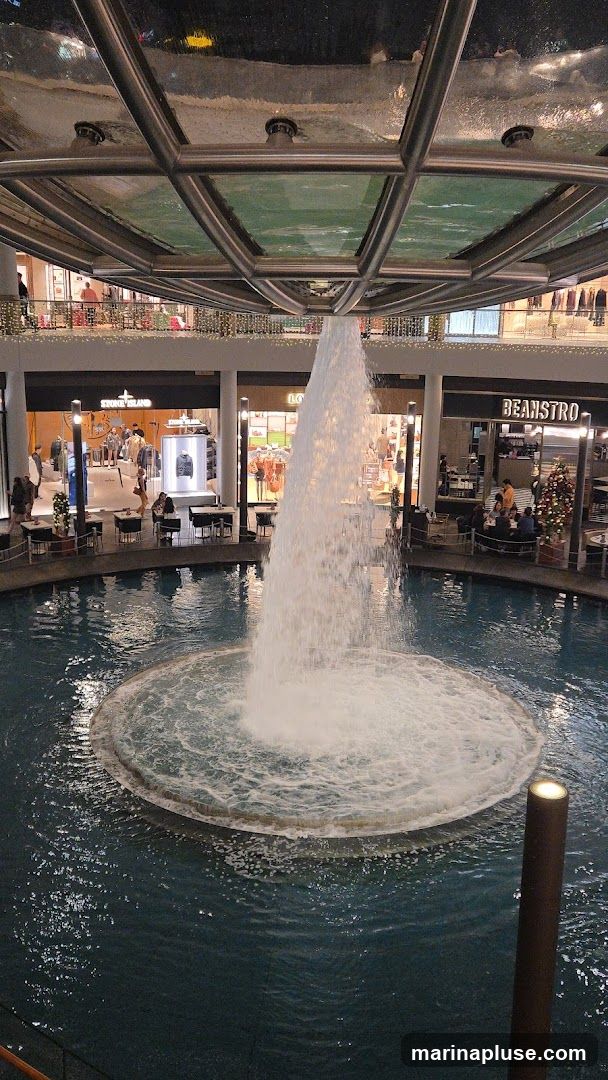 An indoor fountain with a powerful water jet is the central feature of this scene. Surrounding the fountain are shops and seating areas. One shop has a sign that reads "STONE ISLAND" and displays clothing. Another shop has a sign that reads "BEANSTRO" and appears to be a cafe or restaurant, with a Christmas tree visible outside. The architecture suggests a modern shopping mall with a glass ceiling.