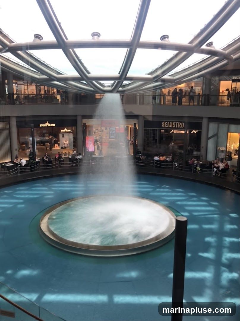 An indoor view of a large circular fountain with water spraying upwards, located in a multi-level shopping mall. Shops with visible signage such as "STONE ISLAND" and "BEANSTRO" are seen on the upper levels surrounding the fountain. People are visible walking and sitting in the mall area.