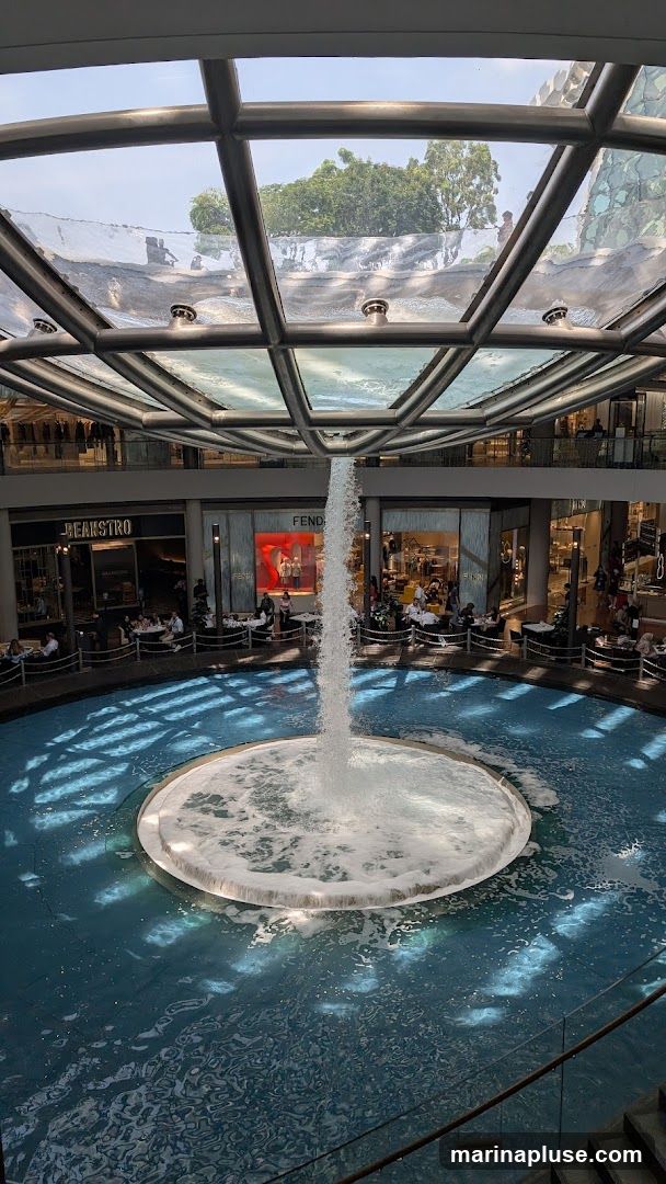 An indoor shopping mall features a large circular fountain with water cascading from above. Surrounding the fountain are multiple shops and restaurants with outdoor seating areas. The architecture includes a modern glass and metal ceiling.