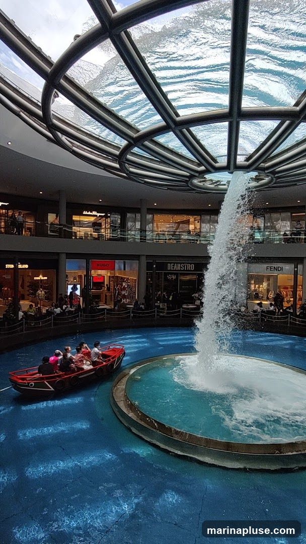 A boat carrying passengers navigates a blue indoor canal beneath a glass ceiling with a waterfall feature. Shops like Salvatore Ferragamo, Hugo, Stone Island, Beanstro, and Fendi are visible on the upper levels surrounding the canal.
