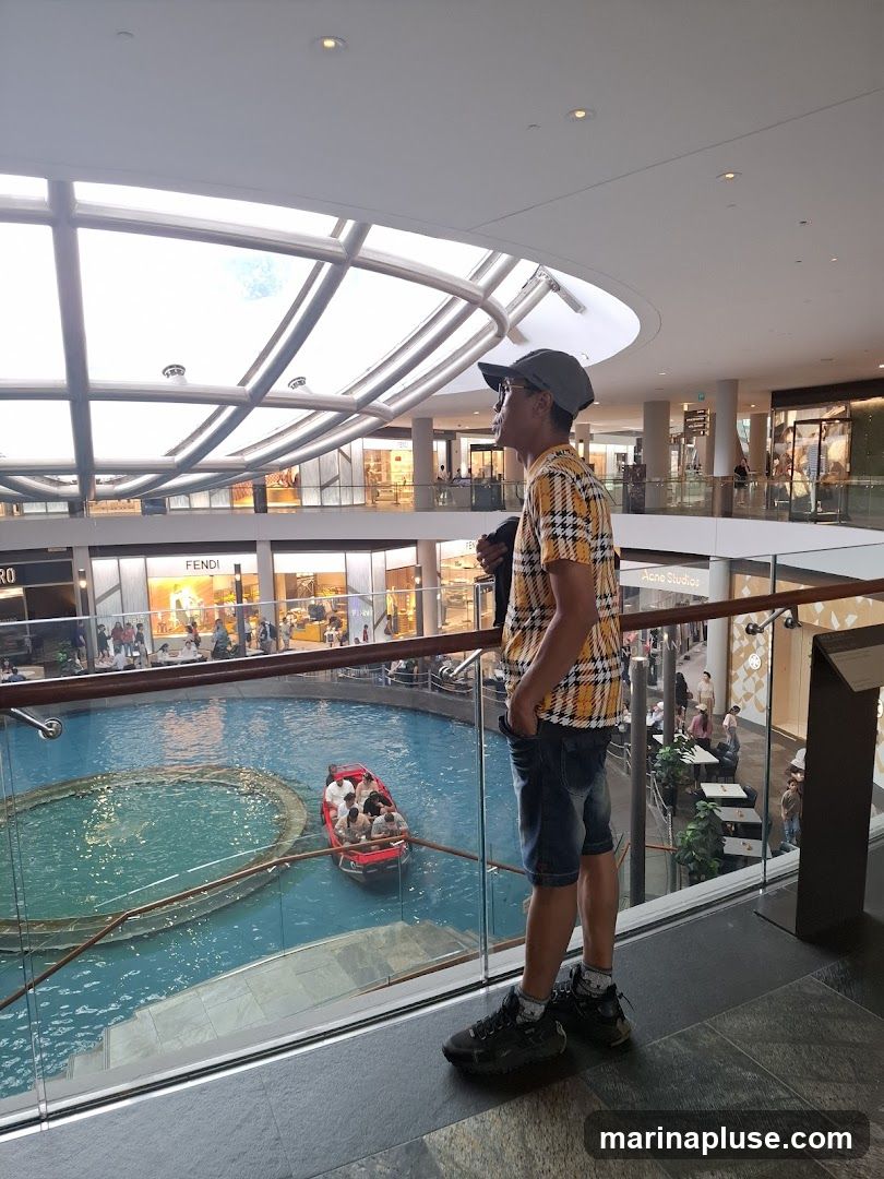 A man stands on a balcony overlooking a large indoor water feature with a boat carrying passengers. The shopping mall has multiple levels with shops visible in the background, including Fendi and Acne Studios. The ceiling is a large glass dome with a metal framework.