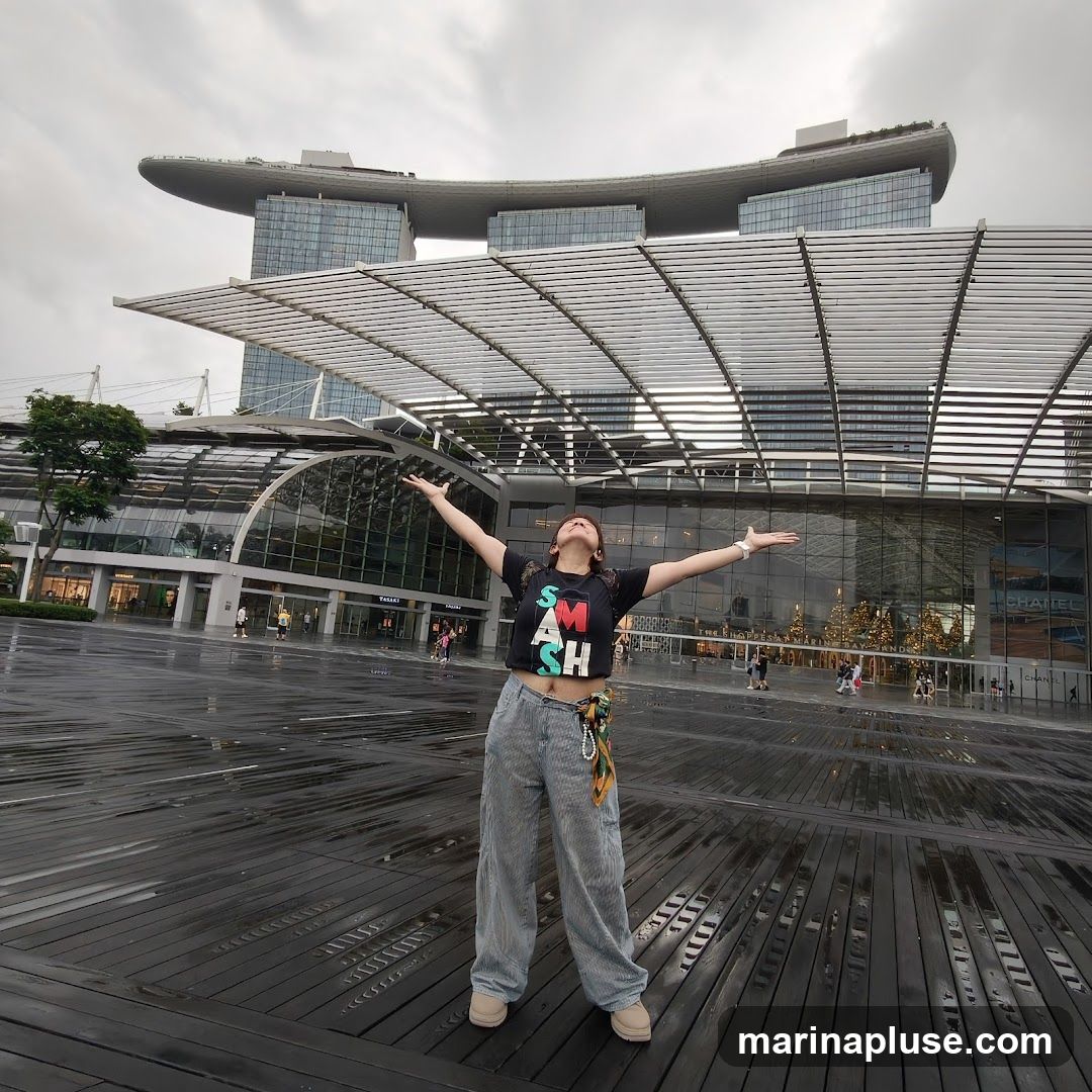 A woman stands with her arms outstretched in front of the Marina Bay Sands hotel in Singapore. The sky is overcast, and the ground is wet, suggesting it may have recently rained. The woman is wearing a black t-shirt with the word "SMASH" printed on it in colorful letters, and striped pants. In the background, the iconic hotel and its connected convention center are visible, along with some Christmas decorations.